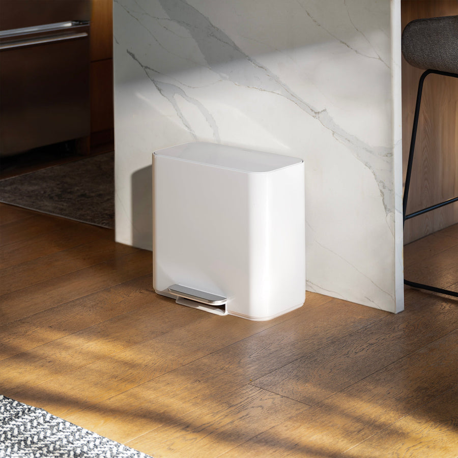 Simplehuman 16L White Mini Step Can with foot pedal, positioned on wood flooring next to a black storage cabinet with books next to it and plants on the other side.
