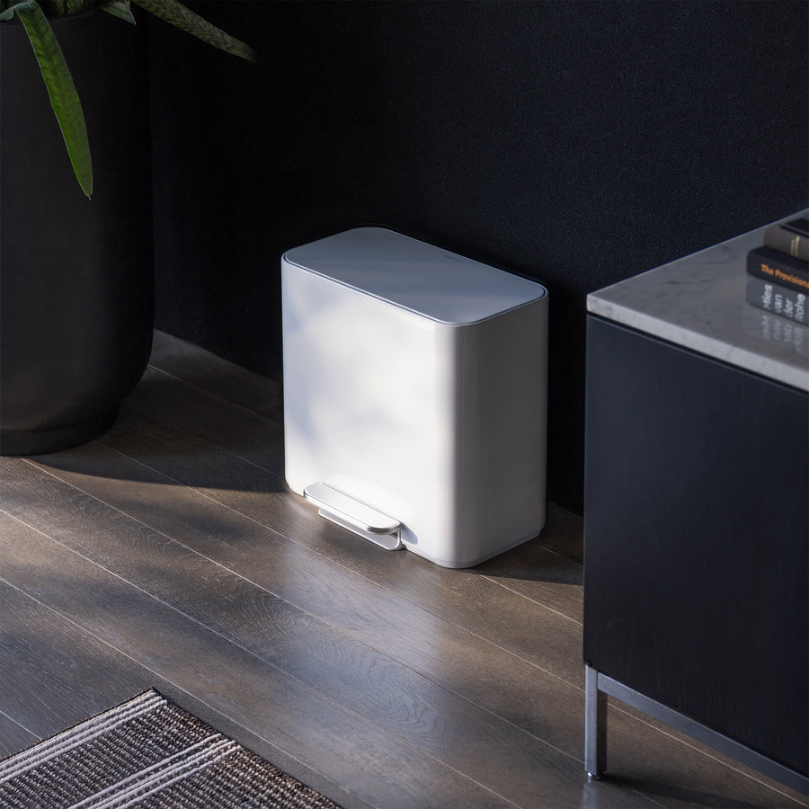 Simplehuman 16L White Mini Step Can with foot pedal, positioned on wood flooring next to a black storage cabinet with books next to it and plants on the other side.