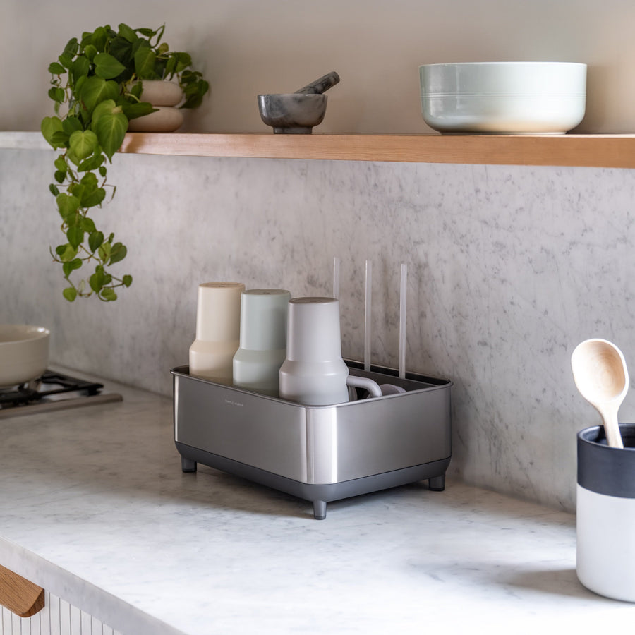 Simplehuman Brushed stainless steel Bottle Dock on marble kitchen countertop holding bottles and mugs beside a plant and utensil holder.