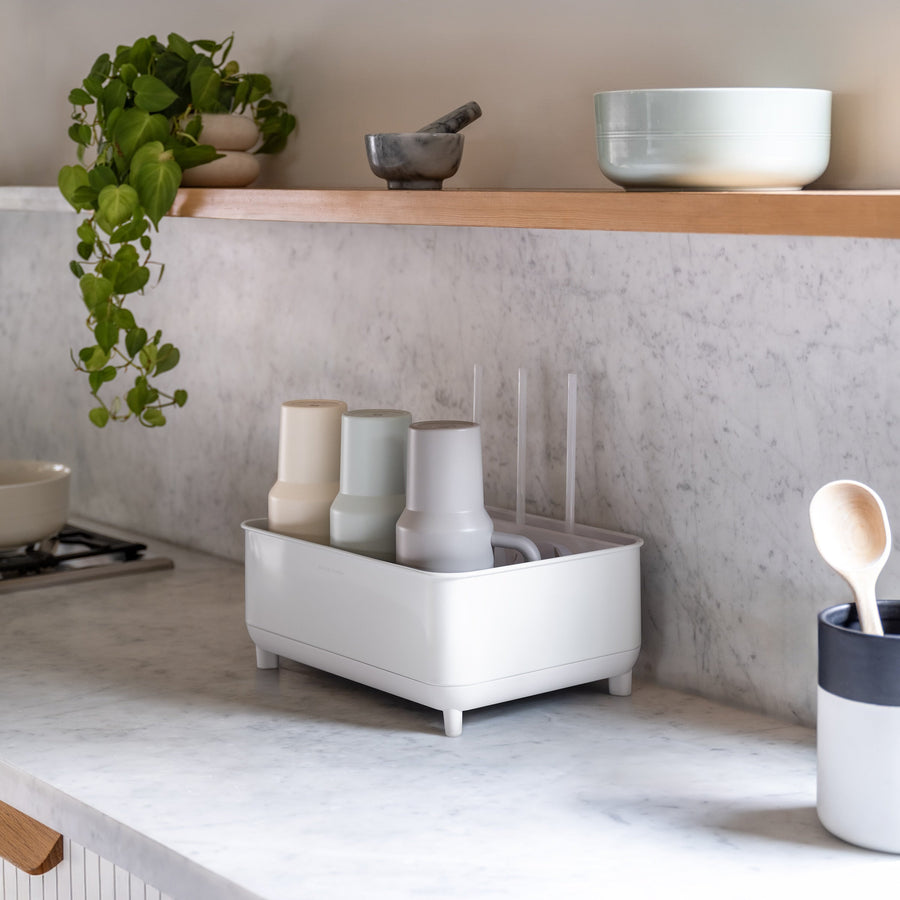 Simplehuman White Bottle Dock on marble kitchen countertop holding bottles and mugs beside a plant and utensil holder.
