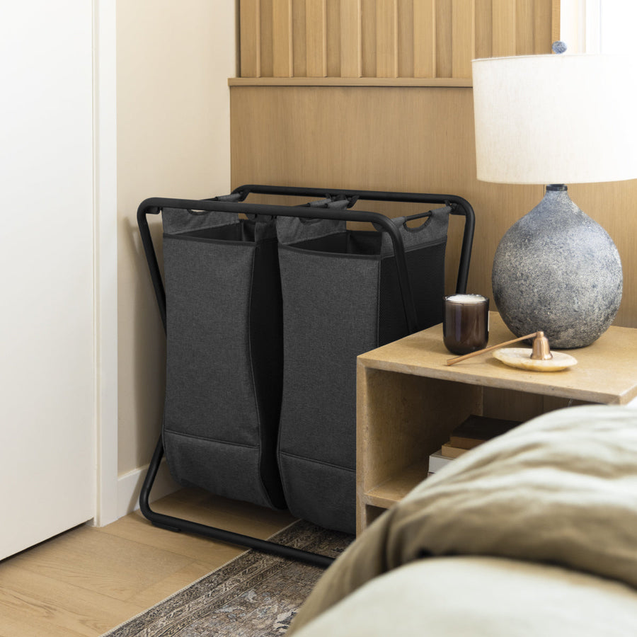 Simplehuman Double X-Frame Hamper with Black frame and dark grey removable liners, ideal for sorting laundry in a bedroom.