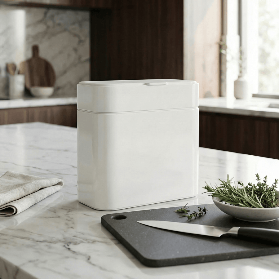 Simplehuman's Compost Caddy in White on white marble countertop with rosemary and thyme on a cutting board and wooden appliances in the background.
